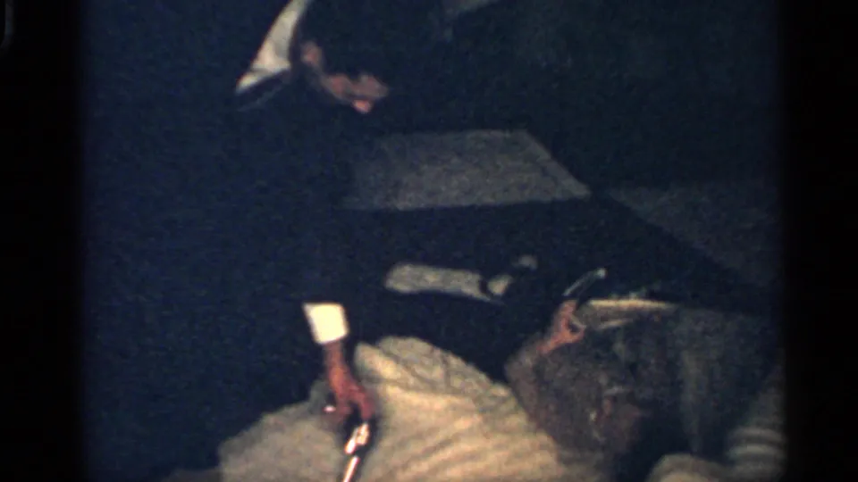 In a dark 1959 Indiana interior, the boy remains on the bed while adult figures stand close by and a small object is visible near him.