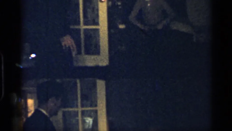 A man in a dark suit stands near a glass-paned door in the dim Indiana home interior, with another figure partly visible at the edge.