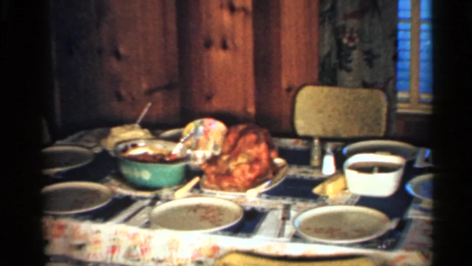 A closer home-movie view of the Indiana dinner table shows a roasted turkey, side dishes, plates, chairs, wood paneling, and a window nearby.