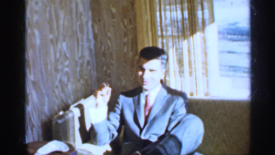 A suited man sits beside a curtained window in a wood-paneled Indiana interior, filmed with bright exposure in 1959 8mm footage.