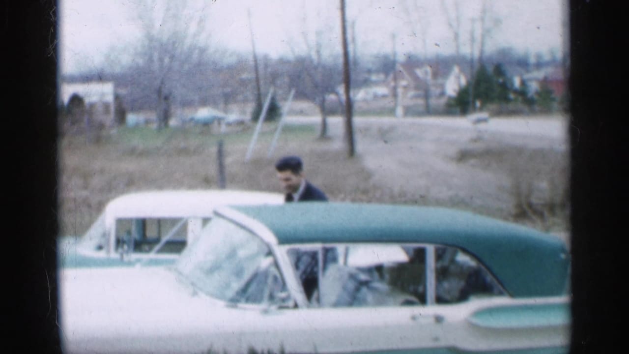Man in a dark suit stands beside a white and teal convertible on a suburban street.