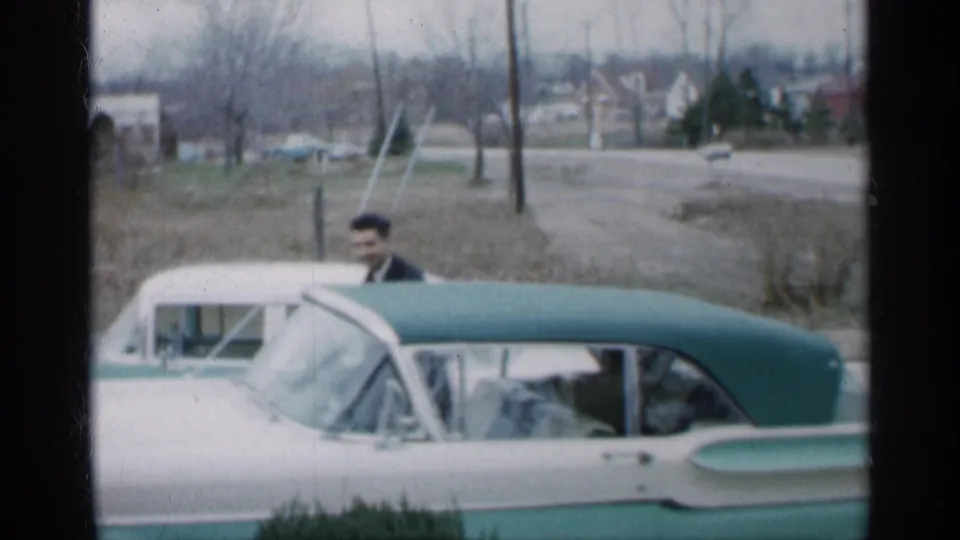 A suited man stands by the two-tone convertible in a residential Indiana setting, with bare trees and modest houses beyond the road.