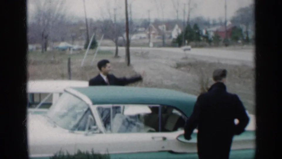 Two suited adults are visible near the two-tone convertible, one gesturing by the car and another partly turned away at the rear.