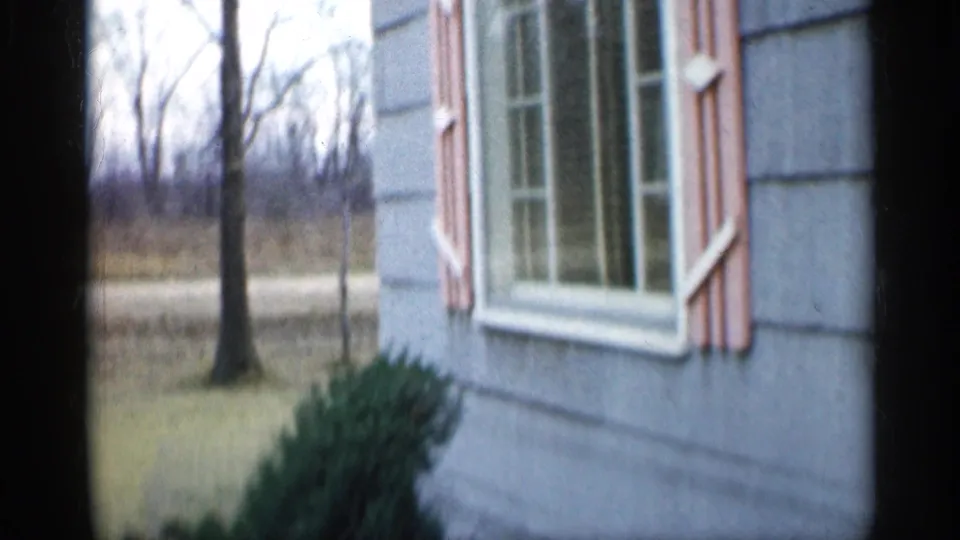 1959 Indiana exterior view of a gray-sided house with pink shutters beside a lawn and bare trees.