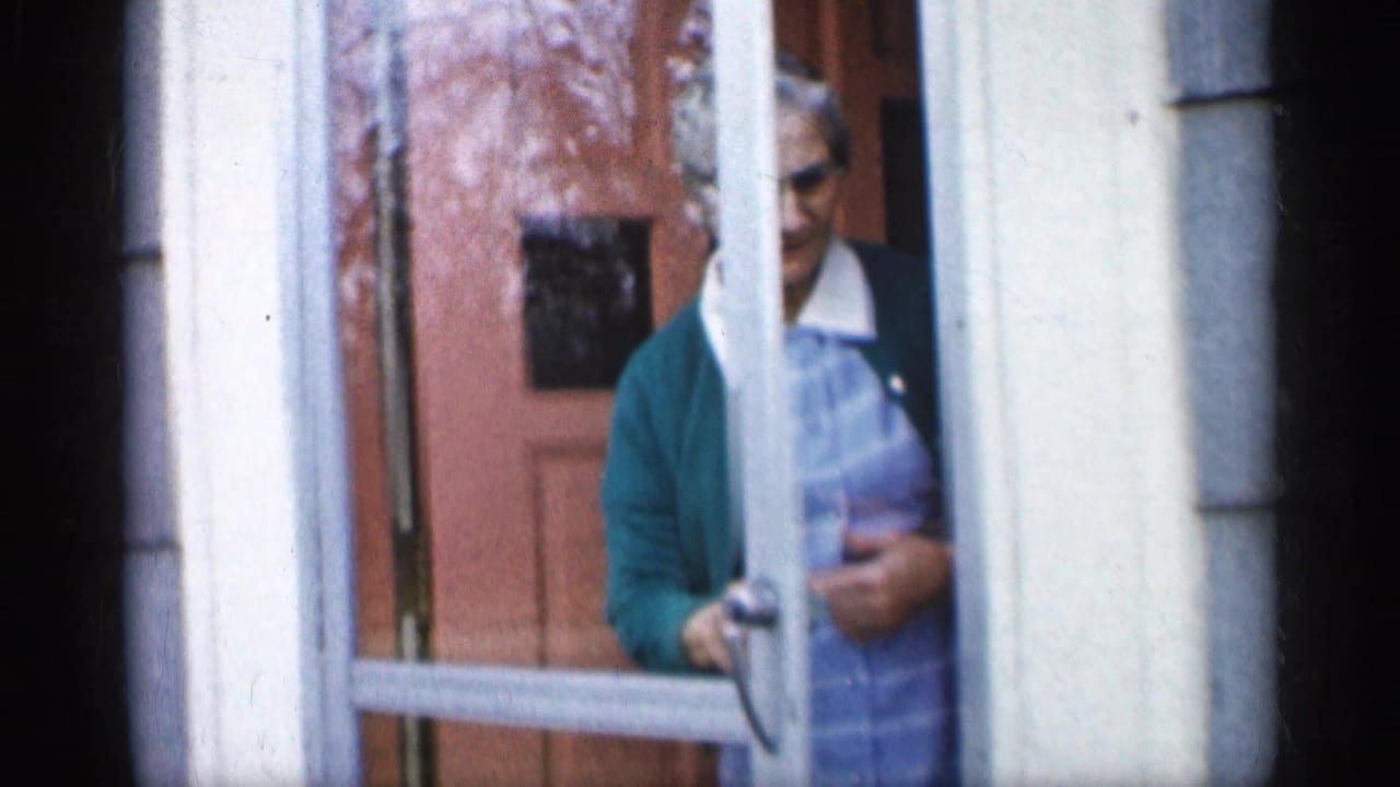In 1959 Indiana home-movie footage, an elderly woman in glasses and a green cardigan stands partly behind a white screen door.