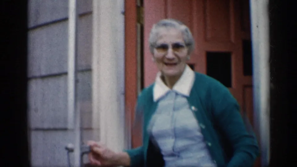 In the Indiana doorway scene, the elderly woman smiles near the open screen door, wearing glasses, a green cardigan, and striped dress.