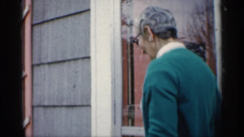 The woman turns away beside the doorway, showing her short gray hair, glasses, green cardigan, and the red door in the background.