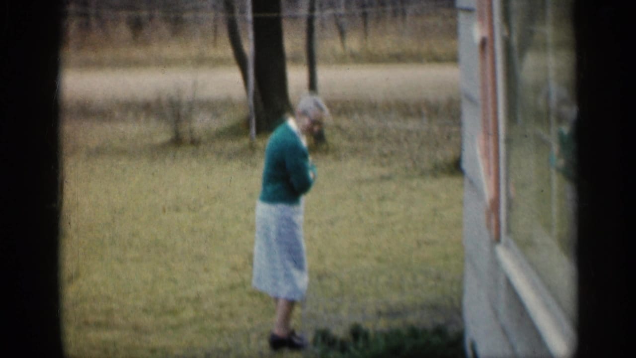 Woman in a green sweater walking on grass beside a house and trees.