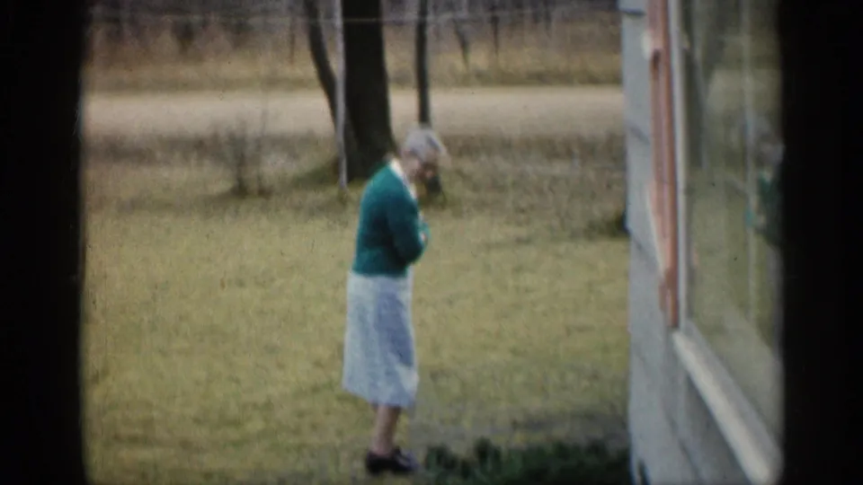 In 1959 Indiana home-movie footage, a woman in a green sweater and light skirt walks across a grassy yard near a house window and bare trees.