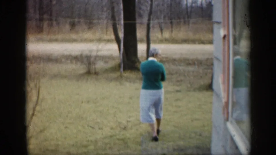 A woman walks away from the camera across a grassy Indiana yard, with bare trees, a roadside, and the side of a house visible nearby.