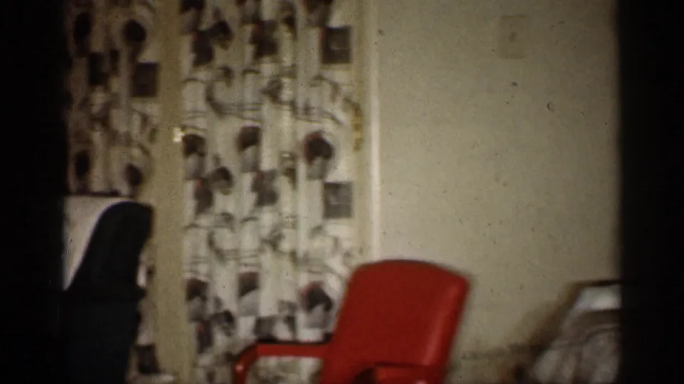 1959 Indiana home-movie frame showing a mid-century living room with patterned drapes, a red chair, and part of a green armchair.