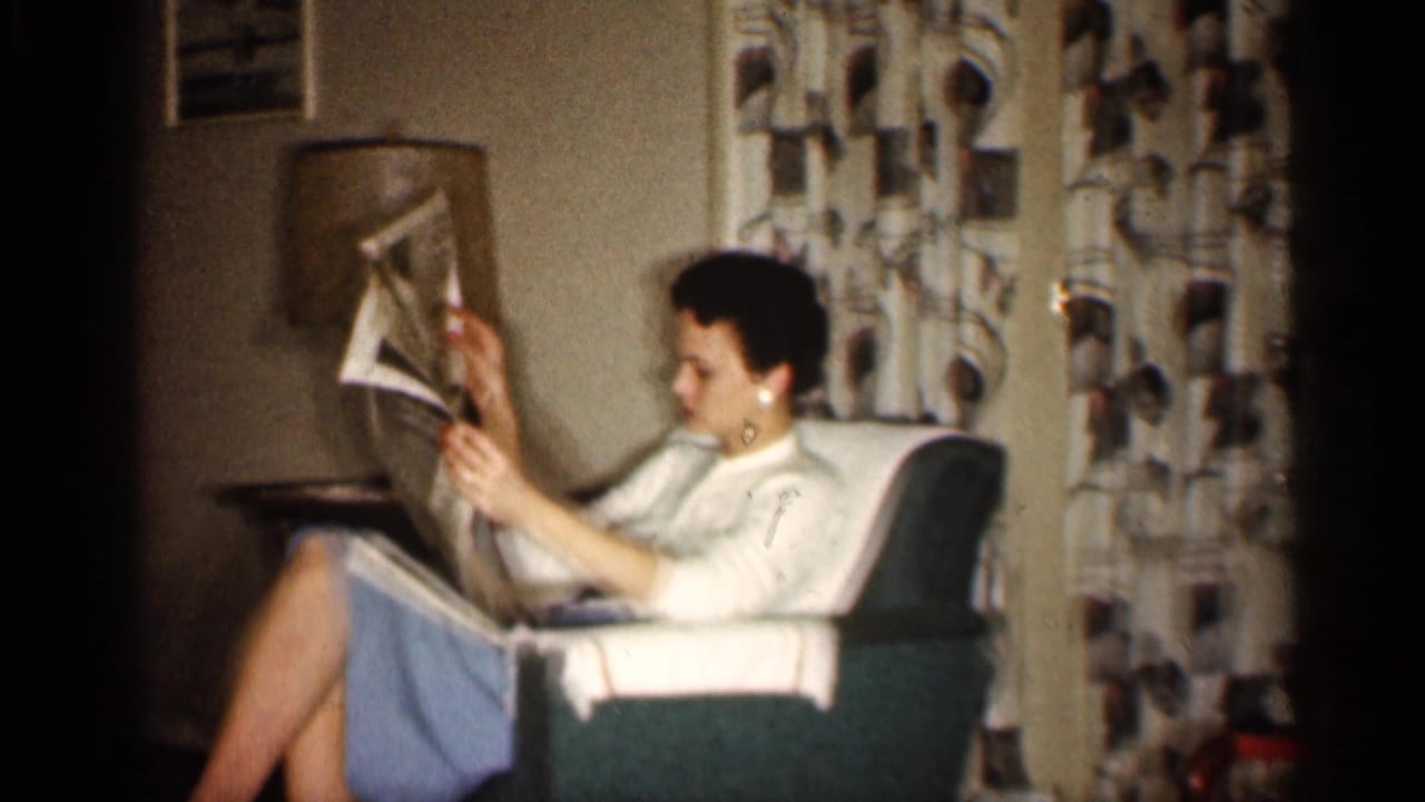 A woman sits in a green armchair in a 1959 Indiana living room, reading a newspaper beside patterned drapes and a lamp.