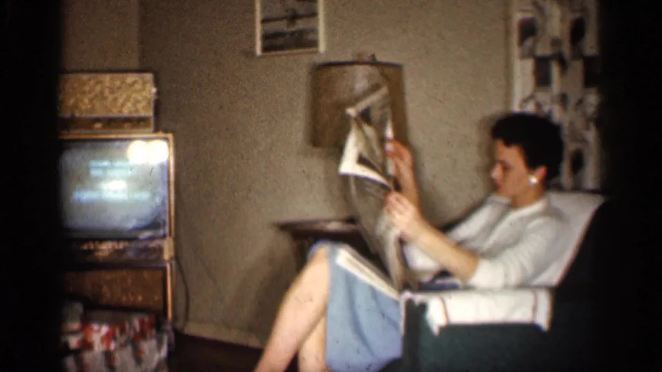 In a 1959 Indiana living room, a woman reads a newspaper while a small television set, lamp, and patterned drapes are visible.
