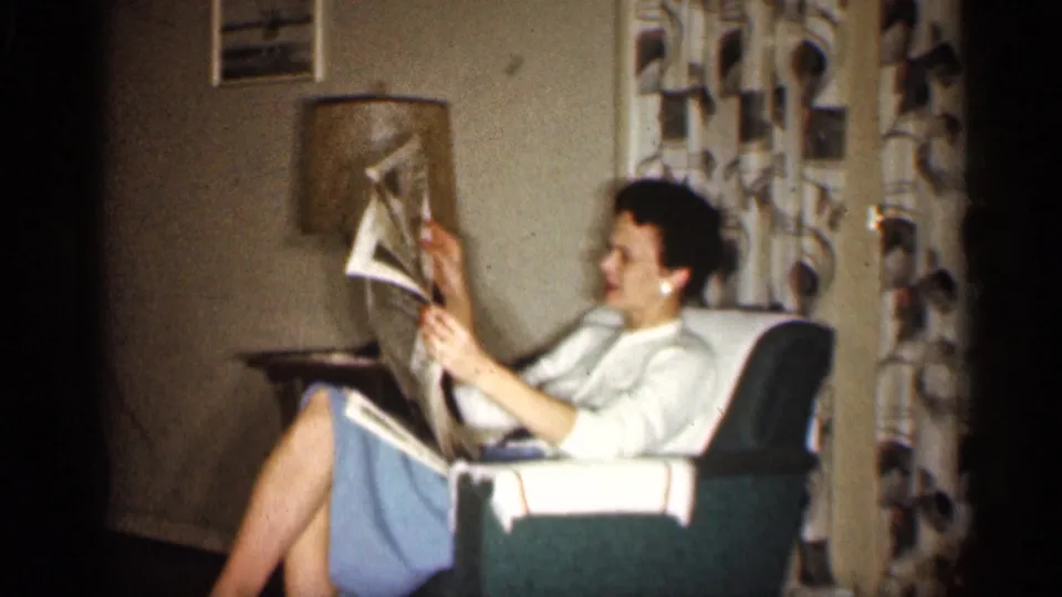 A profile view shows the woman reading a newspaper in a green armchair in the Indiana living room, with drapes and lamp behind her.