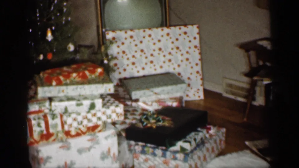 1959 Indiana 8mm frame showing holiday packages by the tree, with a small chair and wooden floor visible in the room.