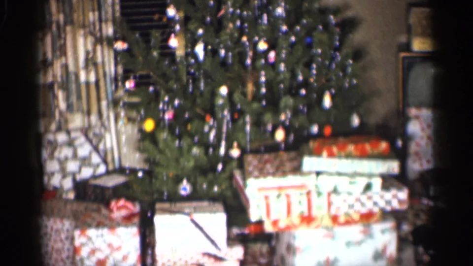1959 Indiana home-movie frame showing a lit Christmas tree, tinsel, ornaments and wrapped presents in a private living room.