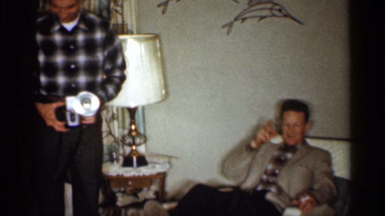 In a 1959 Indiana living room, a man with a handheld movie camera stands near an older man seated by a side table and lamp.