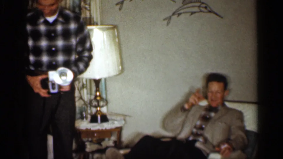 In a 1959 Indiana living room, a man with a handheld movie camera stands near an older man seated by a side table and lamp.