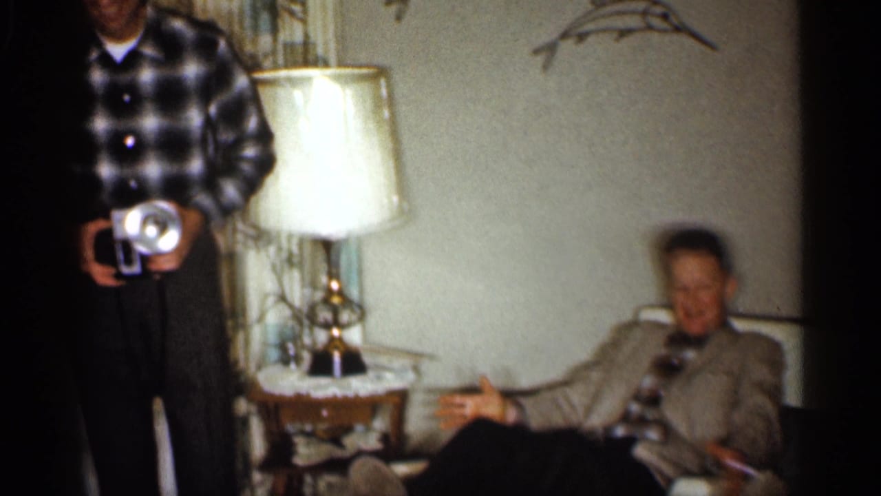 A man in a plaid shirt holds a camera while an older man in a suit gestures from a couch in the Indiana home interior.