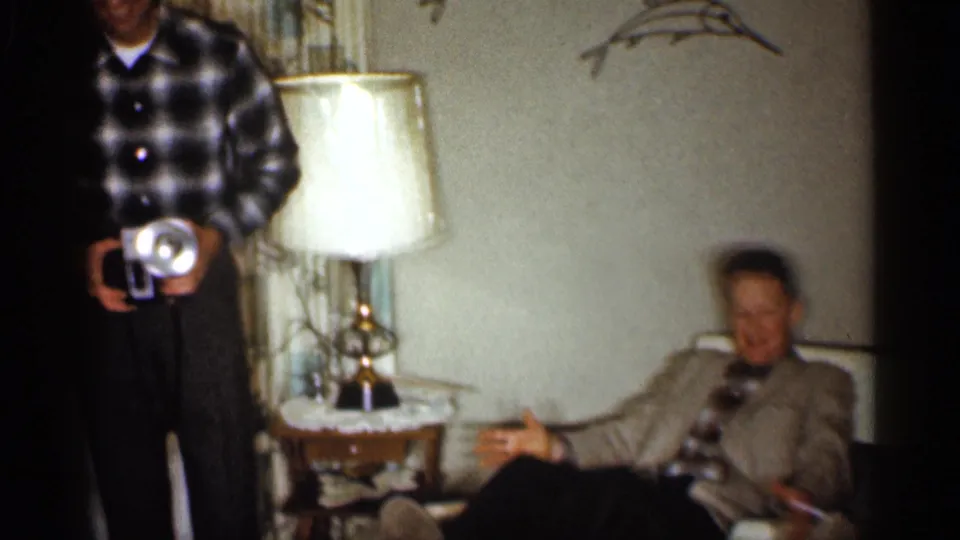 A man in a plaid shirt holds a camera while an older man in a suit gestures from a couch in the Indiana home interior.