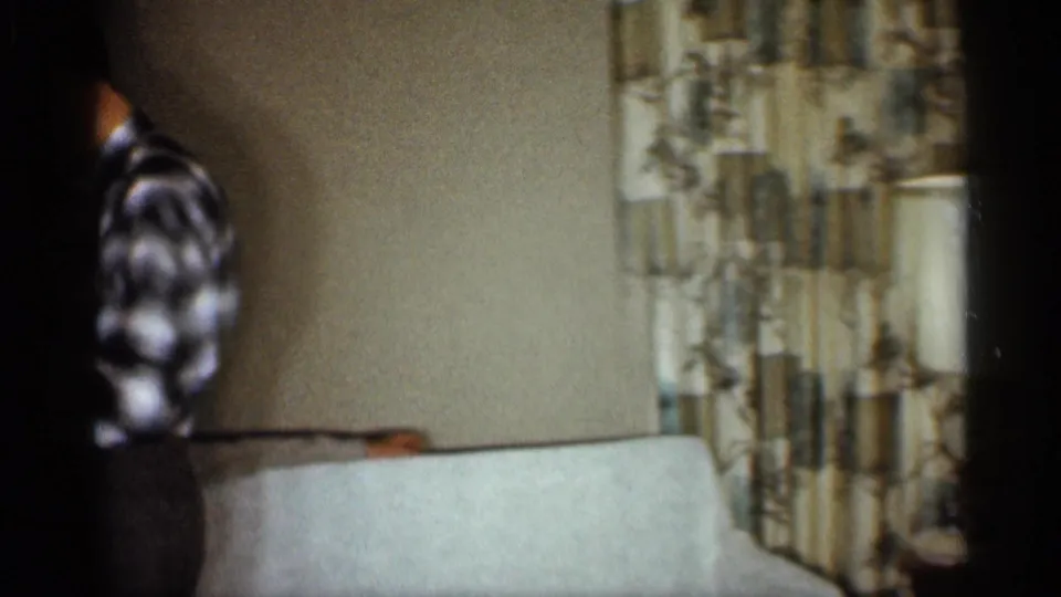 The camera holder turns within the living room, with a couch, patterned curtains, and a lamp visible in the private home setting.