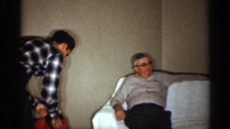 A man bends beside an older man with glasses seated on a couch, continuing the informal 1959 Indiana family-room moment.