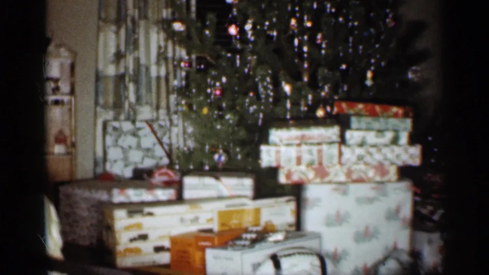An Indiana family Christmas interior shows stacked gift boxes beneath a tinsel-decorated tree, captured on soft 8mm film.