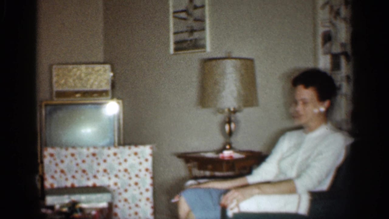 1959 Indiana home-movie frame with a woman seated in a living room near wrapped gifts, a television, lamp, and patterned curtains.