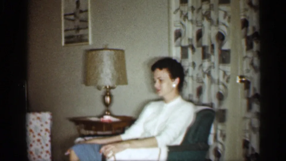 1959 Indiana home-movie frame showing a woman seated in a chair beside a table lamp, wrapped gift edge, and patterned curtains.