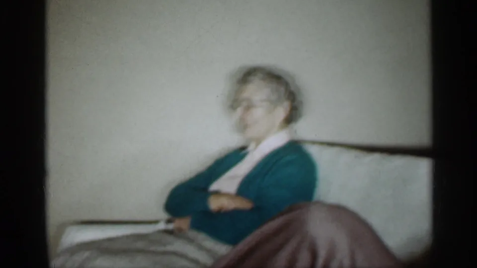 1959 Indiana home-movie frame showing an older man seated on a sofa with arms crossed in a private living room.