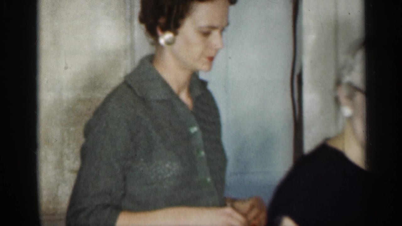 Young woman in a dark collared shirt stands indoors beside an older woman partly visible.