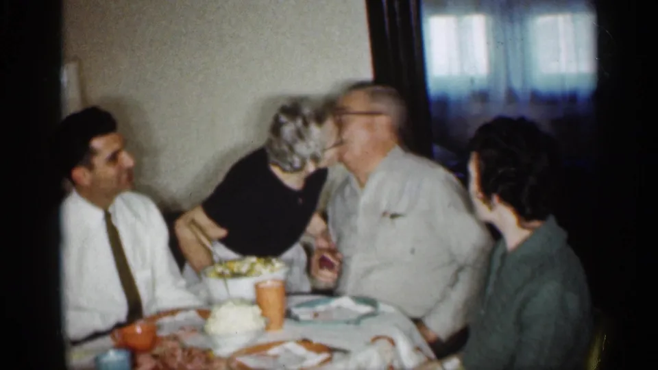 1959 Indiana dining-room frame showing relatives at a table while an older woman leans toward an older man in an affectionate moment.