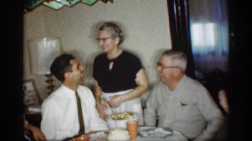 1959 Indiana home-movie frame showing an older woman in glasses standing between two seated men at a family dinner table.