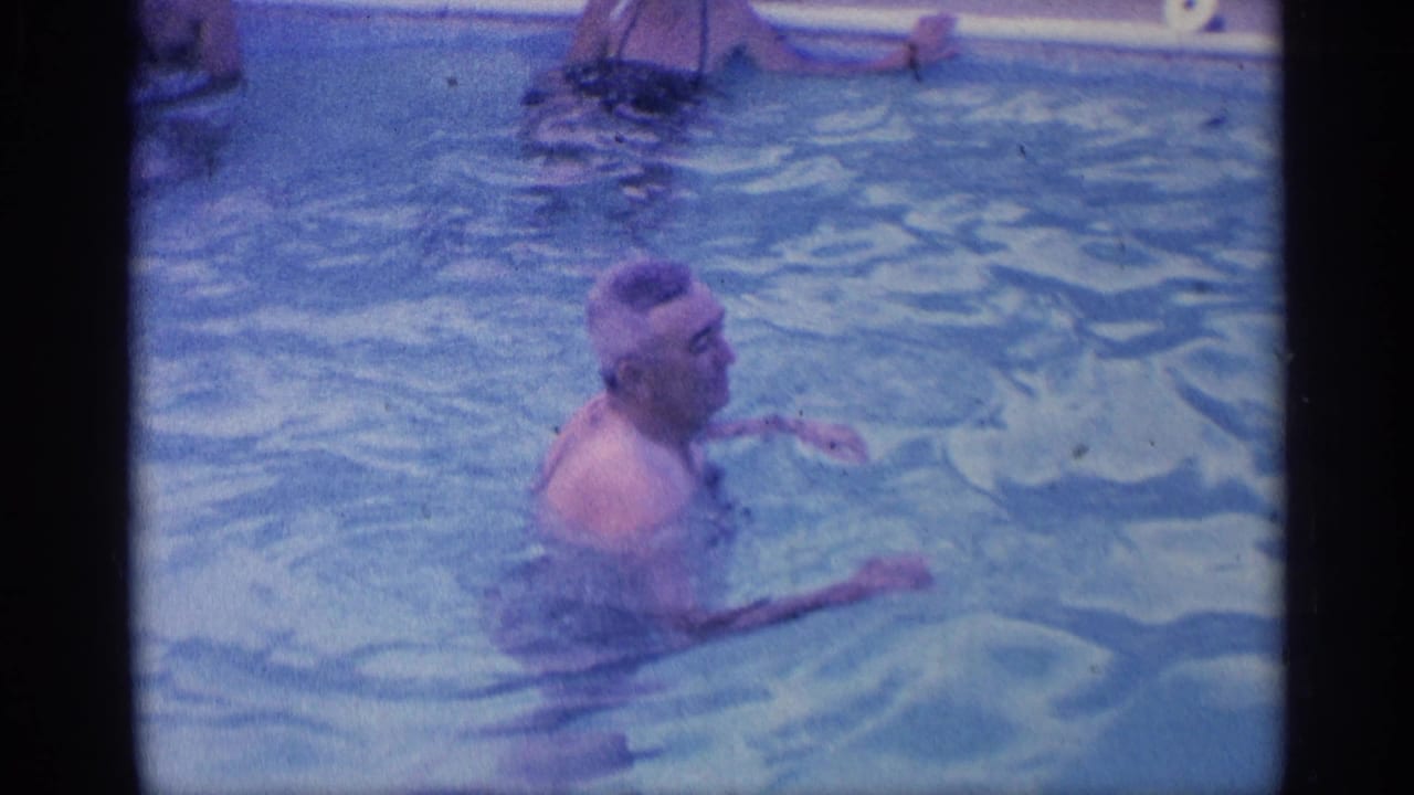 In 1959 Indiana home-movie footage, a man stands chest-deep in a backyard pool as another swimmer is partly visible near the pool edge.