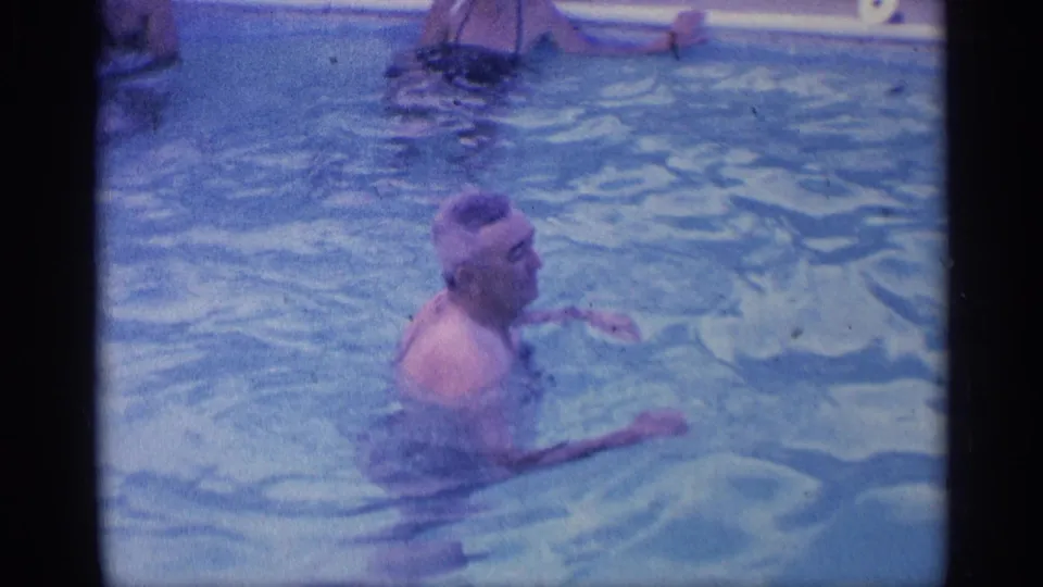 In 1959 Indiana home-movie footage, a man stands chest-deep in a backyard pool as another swimmer is partly visible near the pool edge.