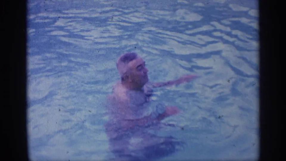A man moves through the backyard pool in this 1959 Indiana home-movie frame, with soft focus, blue tint, and visible 8mm grain.