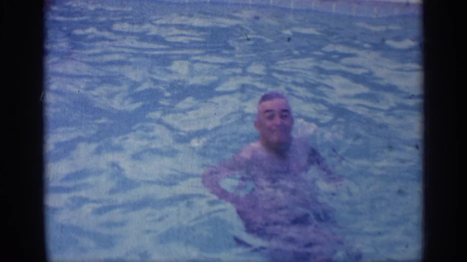The man faces toward the camera from chest-deep pool water in the 1959 Indiana home-movie footage, with visible grain and blue color cast.