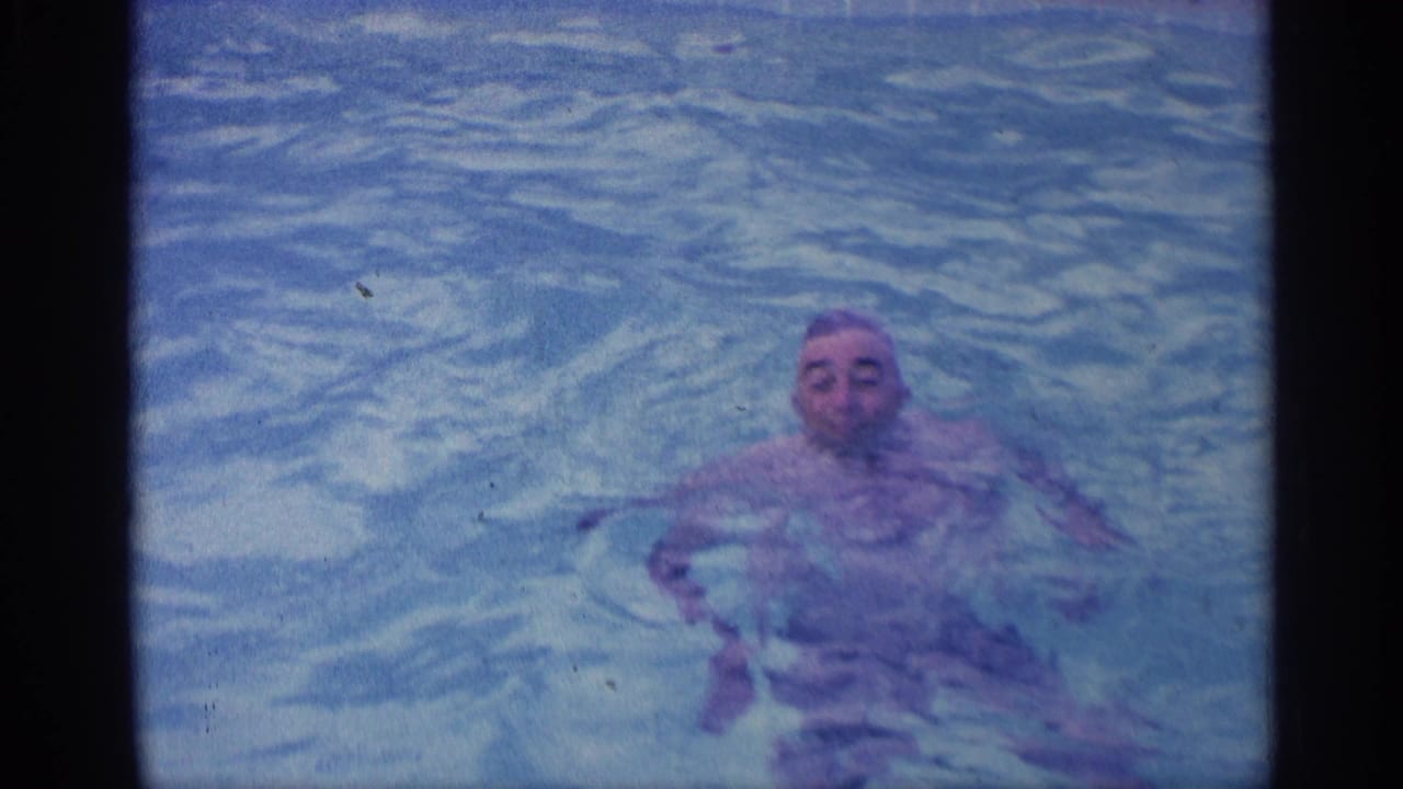 Man floating in a pale blue swimming pool, looking toward the camera.
