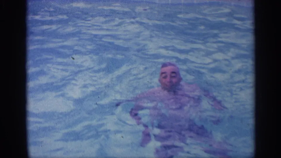 In 1959 Indiana home-movie footage, an adult man floats in a backyard swimming pool with soft blue-purple film color.