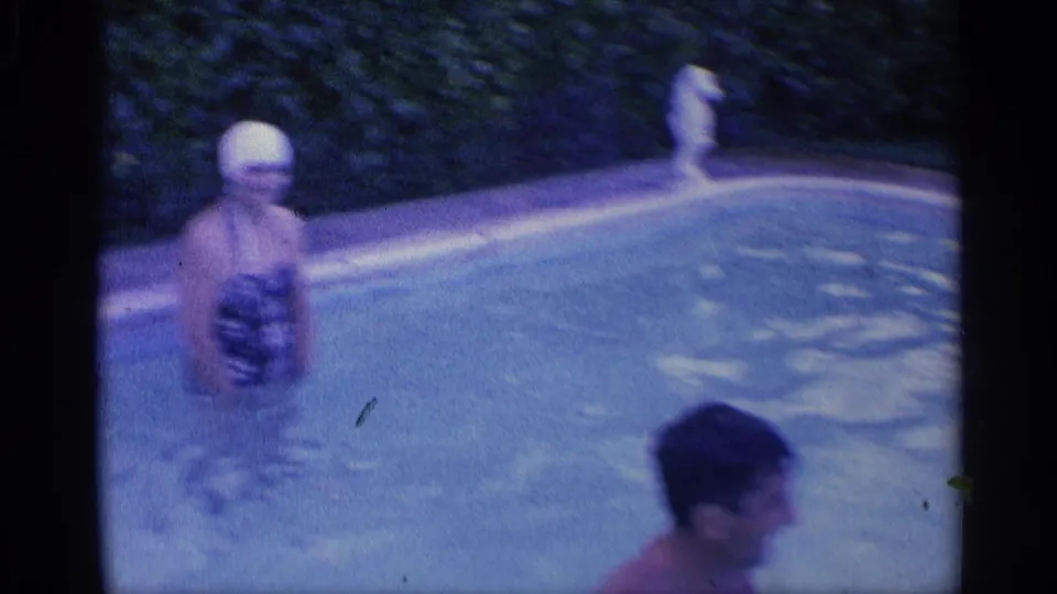 A woman wearing a swim cap stands in the pool while a man moves through the foreground in this 1959 Indiana home movie frame.