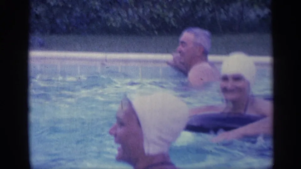 1959 Indiana frame with adult swimmers close to the camera, white bathing caps, blue pool water, and a dark inflatable ring.