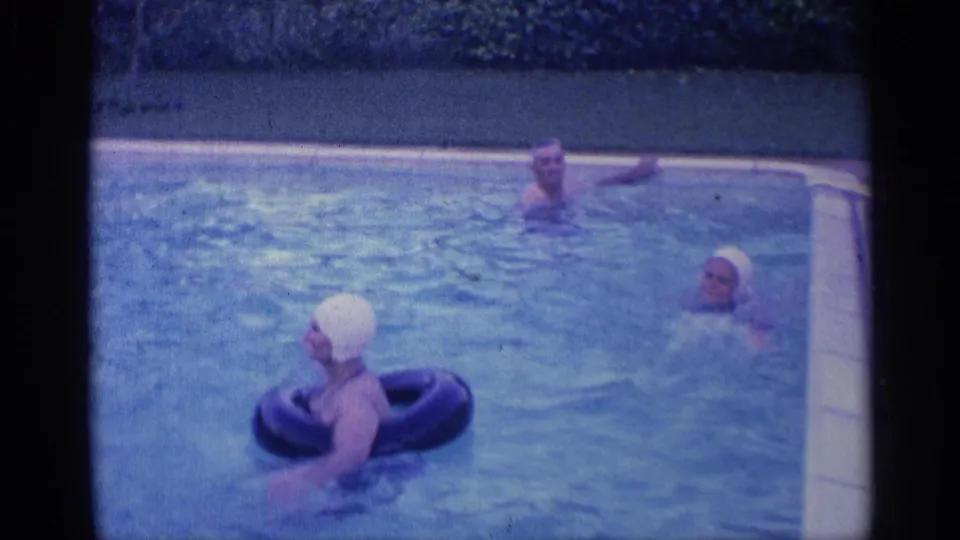 Wide 1959 Indiana home-movie frame showing three adults spread across a backyard pool, with one white-capped swimmer using an inflatable ring.
