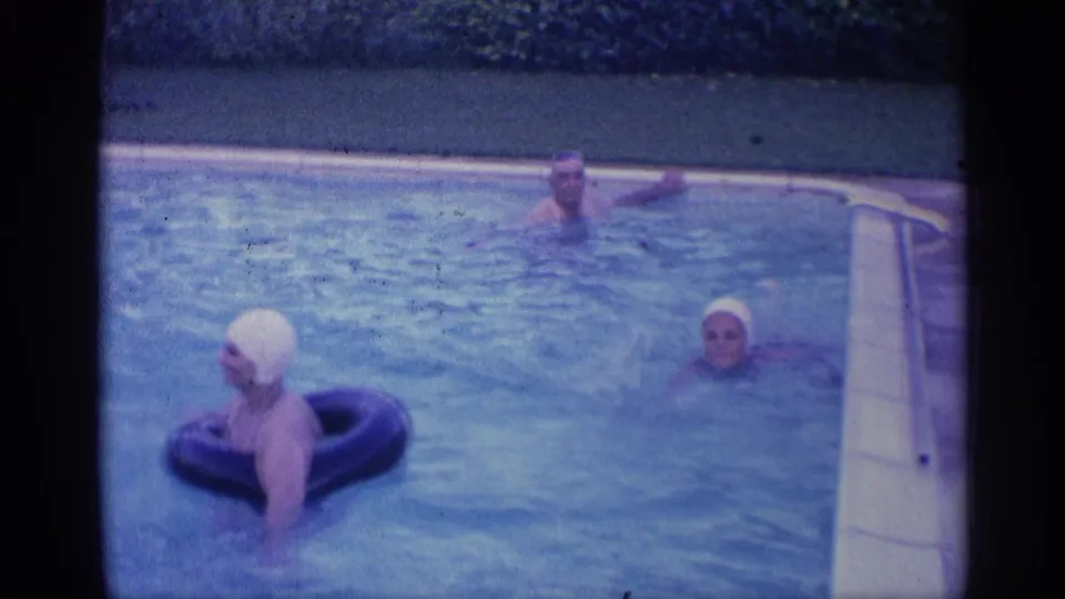 1959 Indiana 8mm frame showing a woman in a white swim cap using a black inflatable ring as two adults swim nearby.