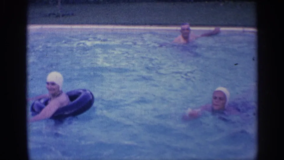 1959 Indiana home-movie frame of two women in white swim caps, with one leaning on a black inflatable ring in a pool.
