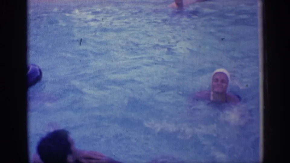 1959 Indiana 8mm frame showing a smiling swimmer in a white cap facing the camera in blue pool water.