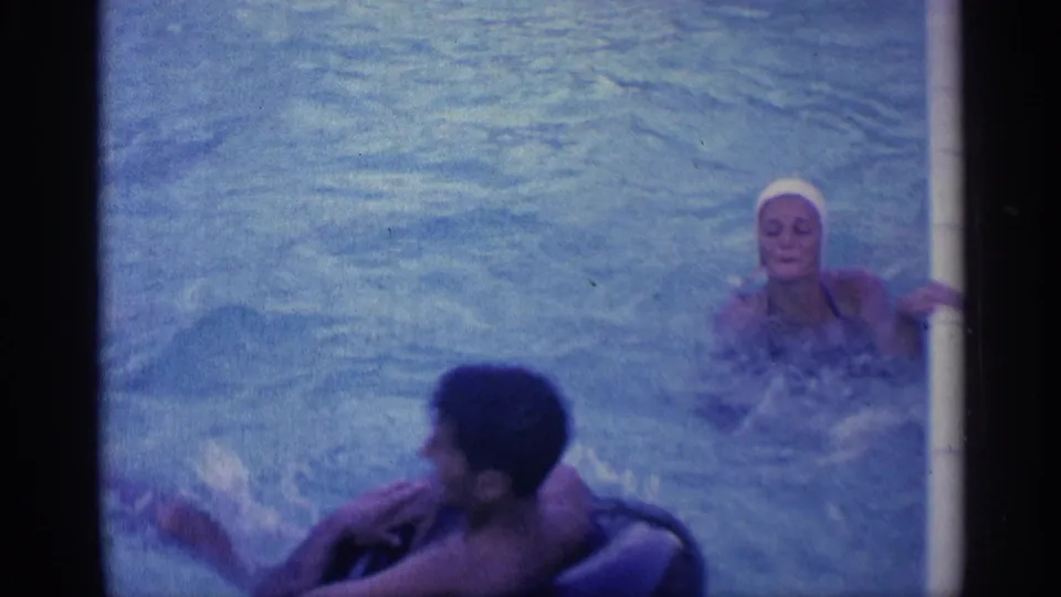 1959 Indiana 8mm frame showing adults in a swimming pool, including a woman in a white swim cap and a man near a dark inner tube.
