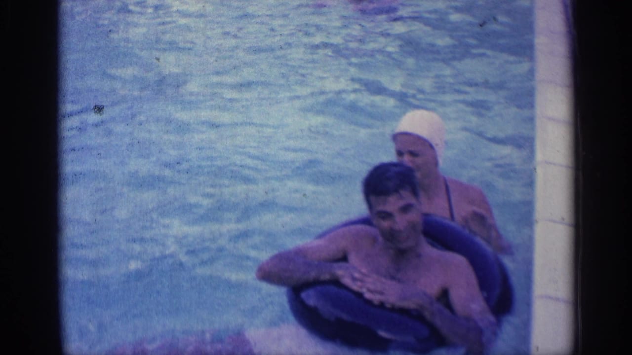 A smiling man leans on a dark inner tube while a woman in a white swim cap swims behind him.