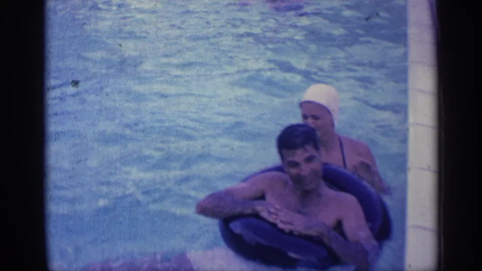 1959 Indiana home-movie frame of a smiling man in a dark inner tube with a woman in a white swim cap nearby in the pool.