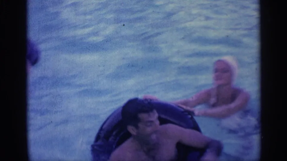 1959 Indiana 8mm frame showing a man partly reclined in a dark inner tube while a woman in a white swim cap swims behind him.