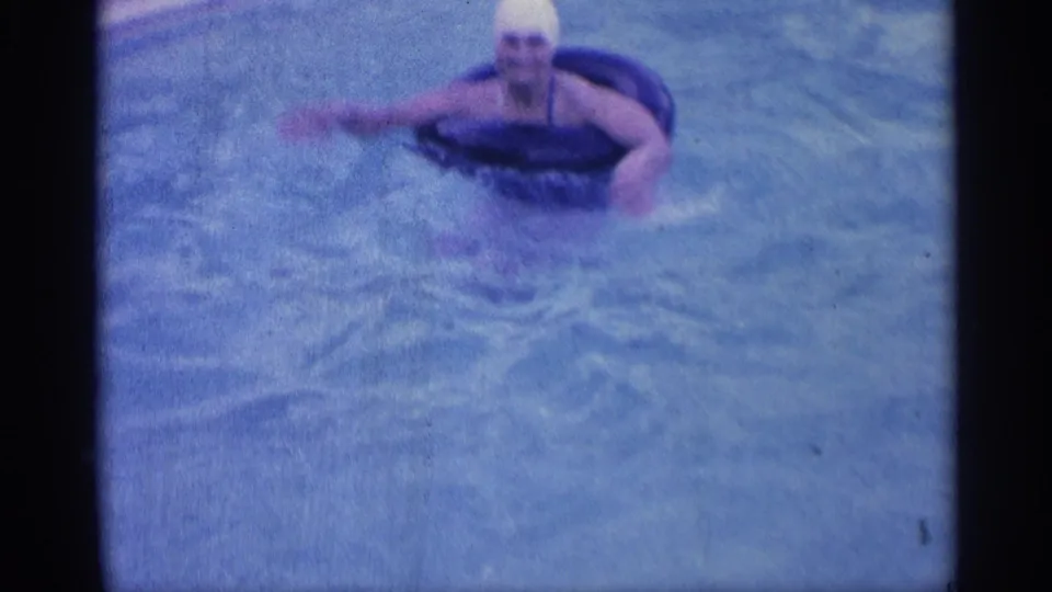 1959 Indiana home-movie frame of a woman in a white swim cap using a dark inner tube while swimming in a pool.